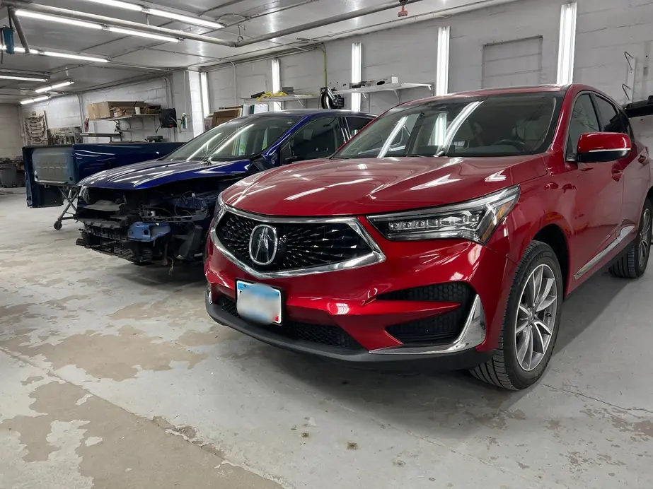 Red Acura RDX in a repair bay beside a blue car under repair